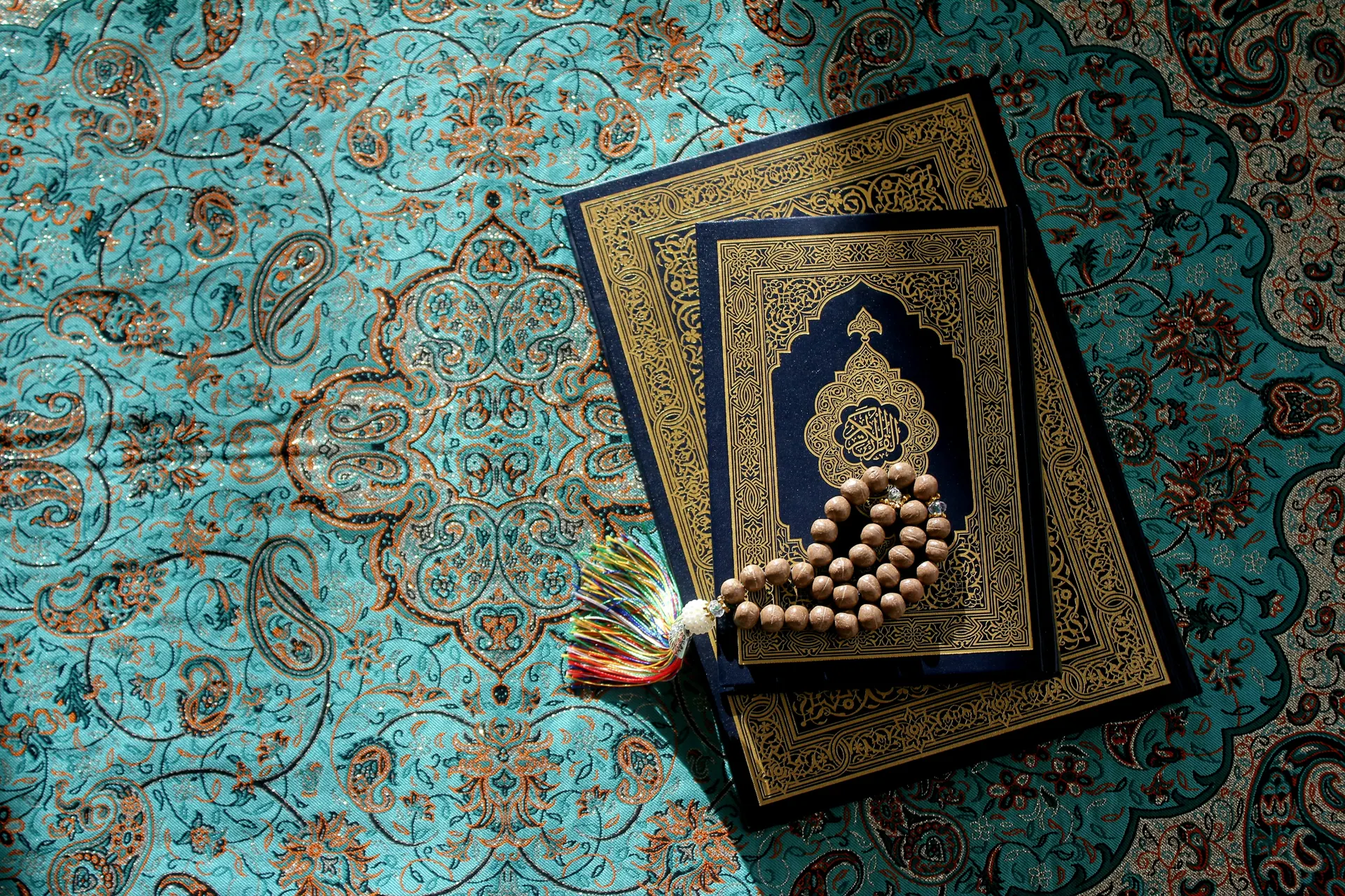 Two ornamented Qurans and a tasbih resting on a teal prayer textile